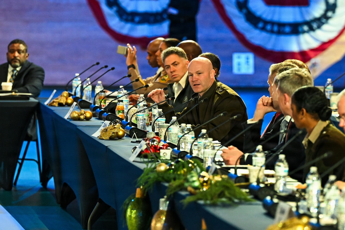 A man wearing a military dress uniform speaks into a microphone while seated at a table. There are other people in business attire and military dress uniforms seated at the table with microphones in front of them. There are water bottles and festive holiday decorations on the table.