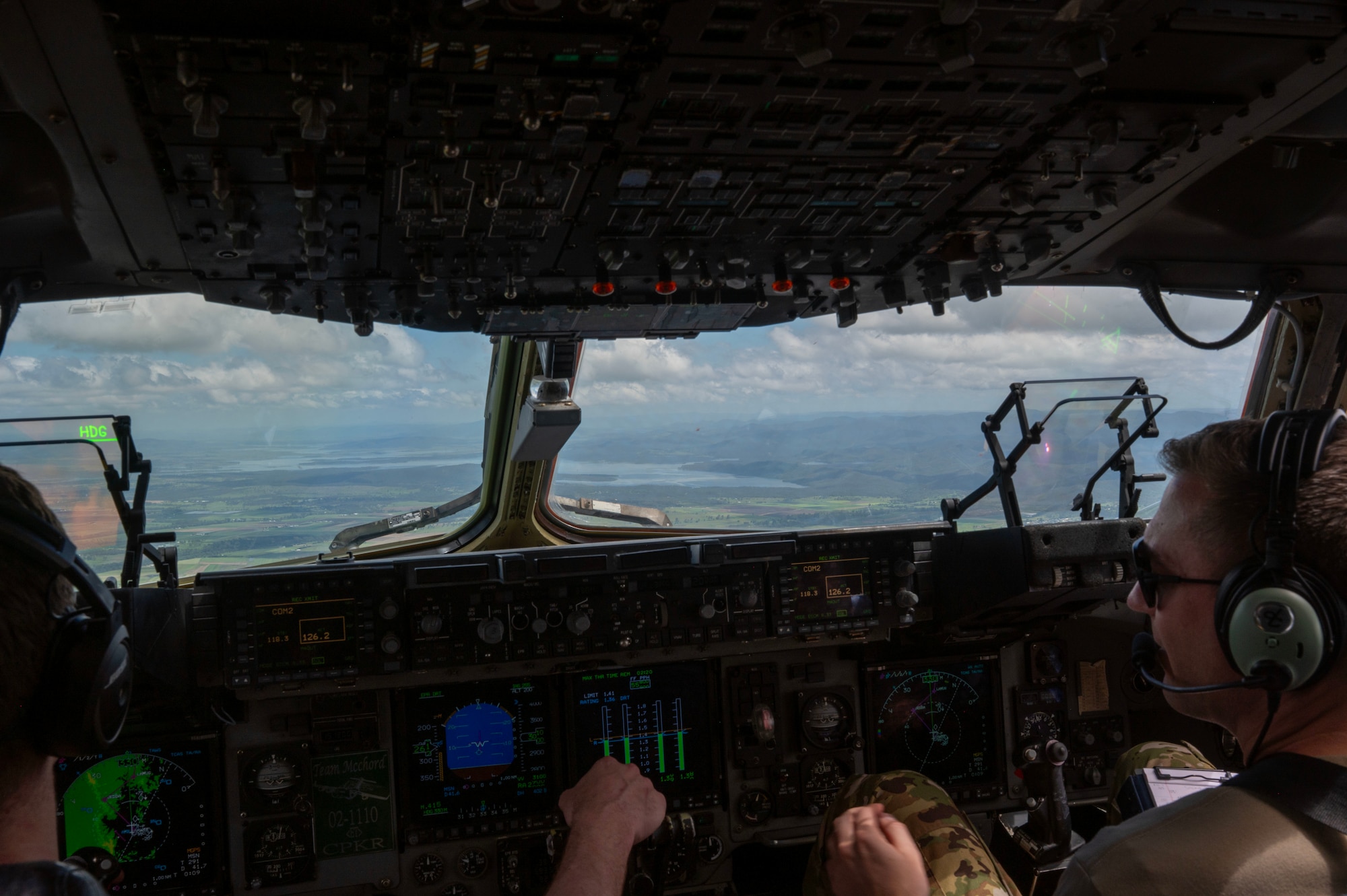 A shot taken from behind two pilots in the cockpit of a cargo plane. A green landscape can be clearly seen through the front windows