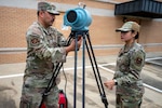Two uniformed service members stand outdoors in front of radiation detection equipment.
