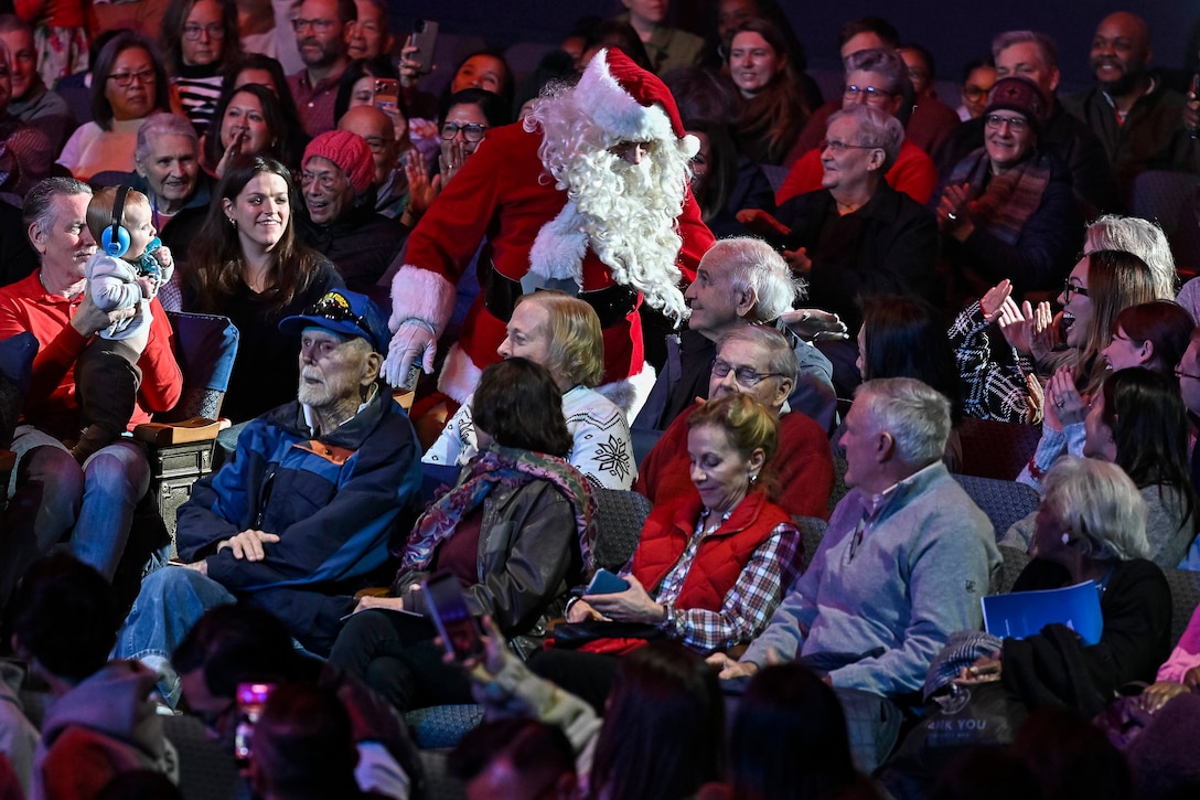 A band member, dressed as Santa, walks through a seated audience in a dimly lit concert hall.