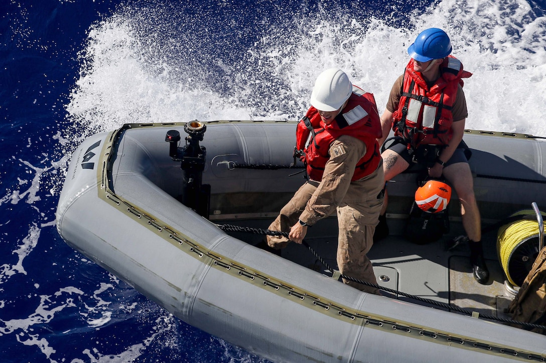 Two sailors wearing hard hats and red life vests ride in a small boat as water splashes behind them.