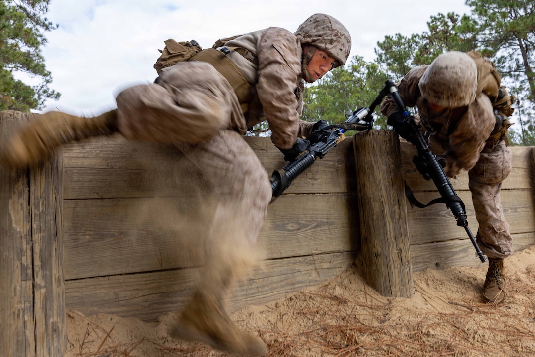Two Marine Corps recruits in tactical gear carrying weapons crawl over an obstacle in a wooded area.