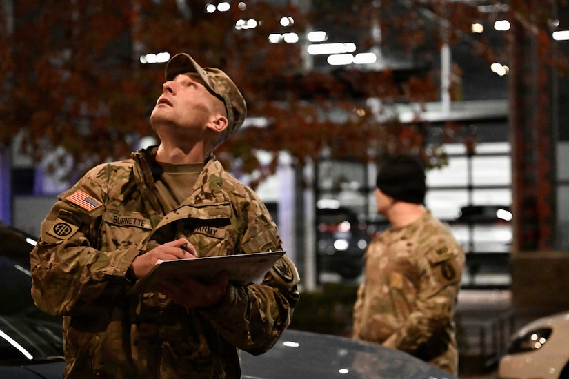 A man in a camouflage military uniform looks up, observing something out of frame while holding a clipboard.