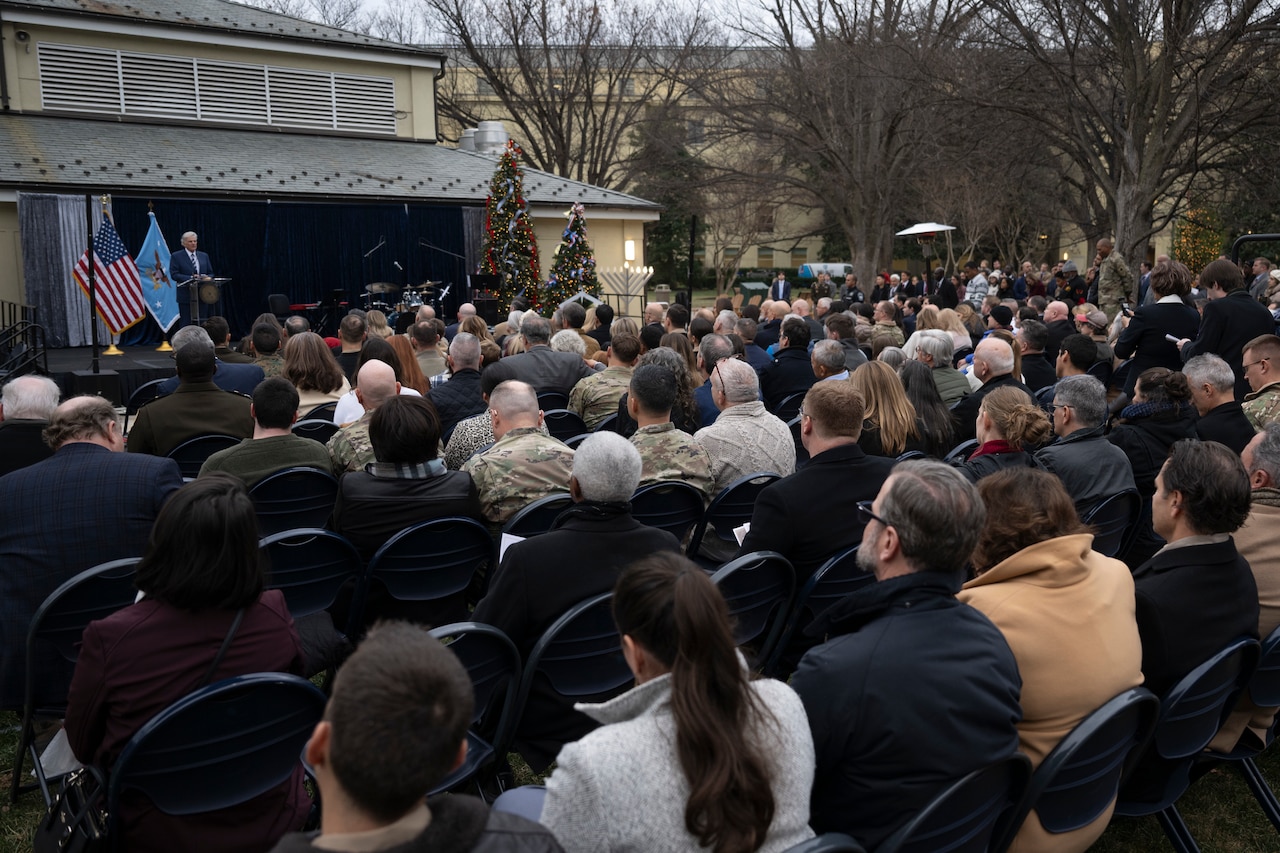 Dozens of personnel, both military and civilian, are seated outdoors looking at a man in a suit speaking behind a lectern.
