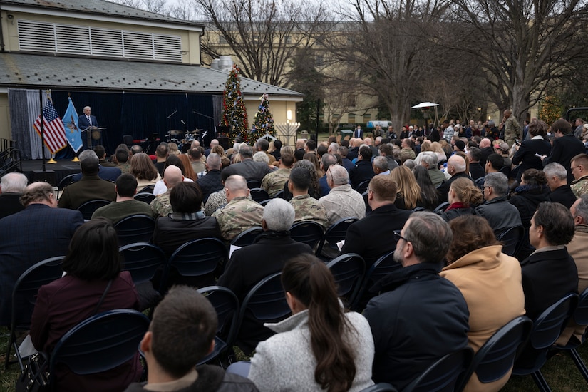 Dozens of personnel, both military and civilian, are seated outdoors looking at a man in a suit speaking behind a lectern.