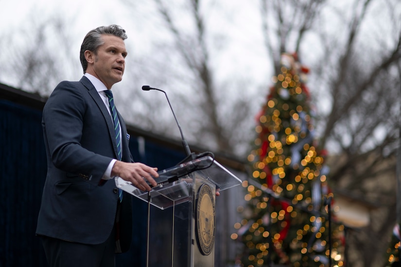 A man in a suit stands behind a lectern. Behind him are out of focus Christmas trees.