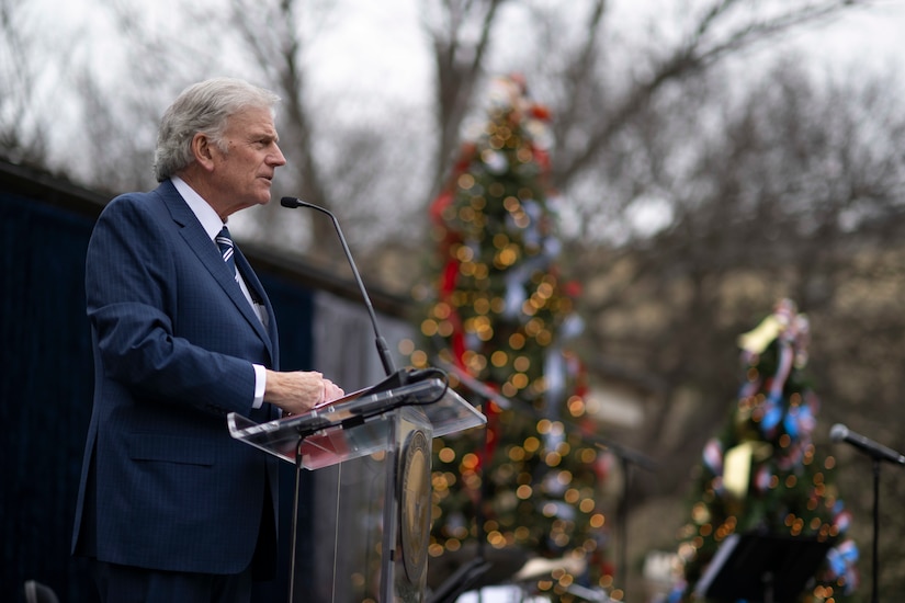 A man in a suit stands behind a lectern. Behind him are out of focus Christmas trees.