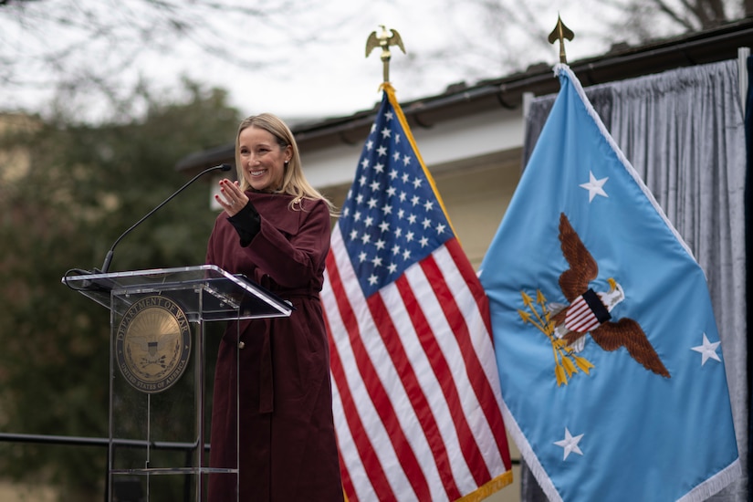 A woman in business attire stands behind a lectern. Behind her are the U.S. and secretary of war flags.
