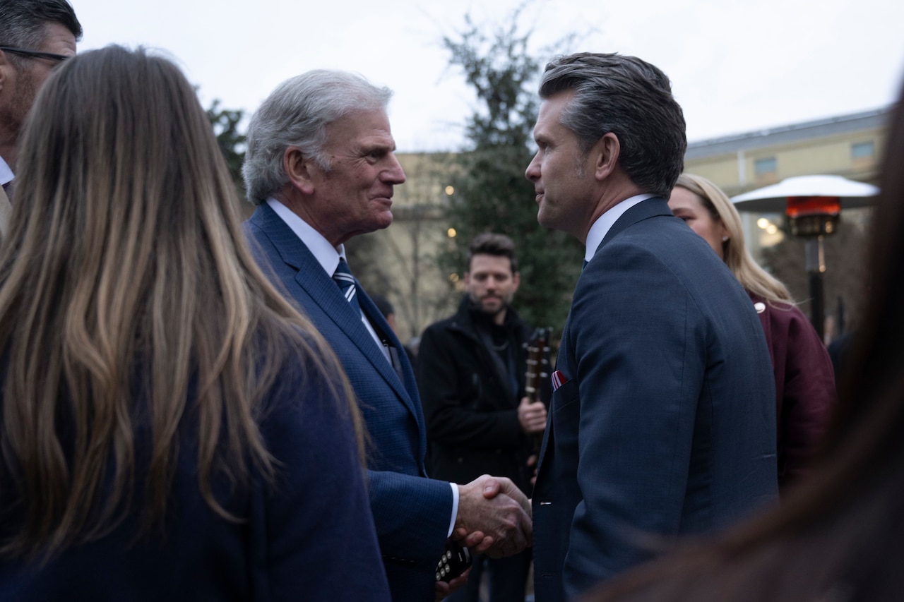 Two men in suits shake hands while other people stand around them.