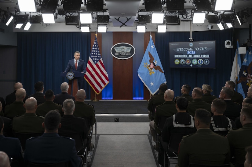 A man in business attire stands behind a lectern speaking into a microphone. Behind the man is an American flag, another flag with a large eagle in the center and a sign depicting a five-sided building that reads, “The Pentagon. A large group of people, their backs to the camera, are listening to the man speak.