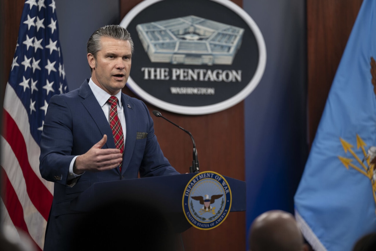 A man in business attire stands behind a lectern speaking into a microphone. A seal on the lectern shows an eagle and reads, “Department of War.” Behind the man is an American flag and a sign depicting a five-sided building that reads, “The Pentagon.”