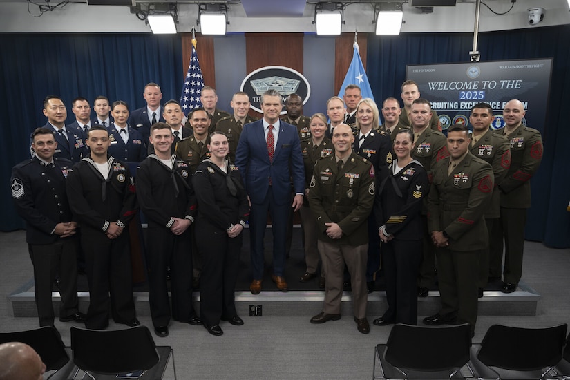 A large group of people in various dress military uniforms pose for a photograph with a man in the center of the group wearing civilian business attire.