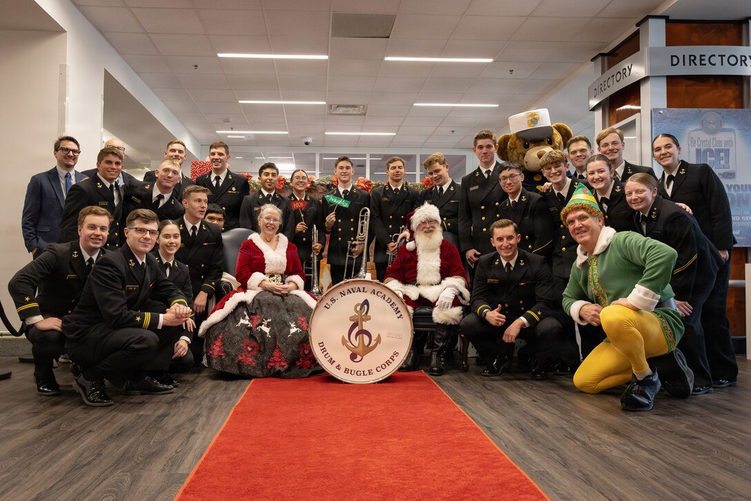 Members of the U.S. Naval Academy Drum and Bugle Corps pose for a group photo with Santa Claus, Mrs. Claus and Buddy the Elf during a Toys for Tots charity event at the Pentagon, Washington, D.C., Dec. 9, 2025. Since 1947, the Marine Corps Reserve has spearheaded the Toys for Tots program nationwide to collect toys for less fortunate children during the holiday season. Presently, the program distributes 7 to 18 million toys to children annually. (U.S. Marine Corps photo by Lance Cpl. Van Hoang)