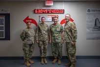 Four people in military uniforms pose in front of a clock.