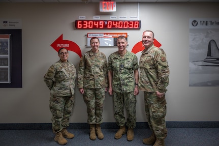 Four people in military uniforms pose in front of a clock.
