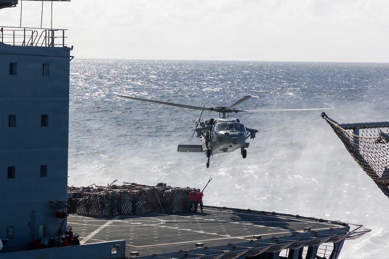 A military helicopter hovers over the deck of a large ship at sea while two people in military uniforms attempt to attach cargo to the aircraft.
