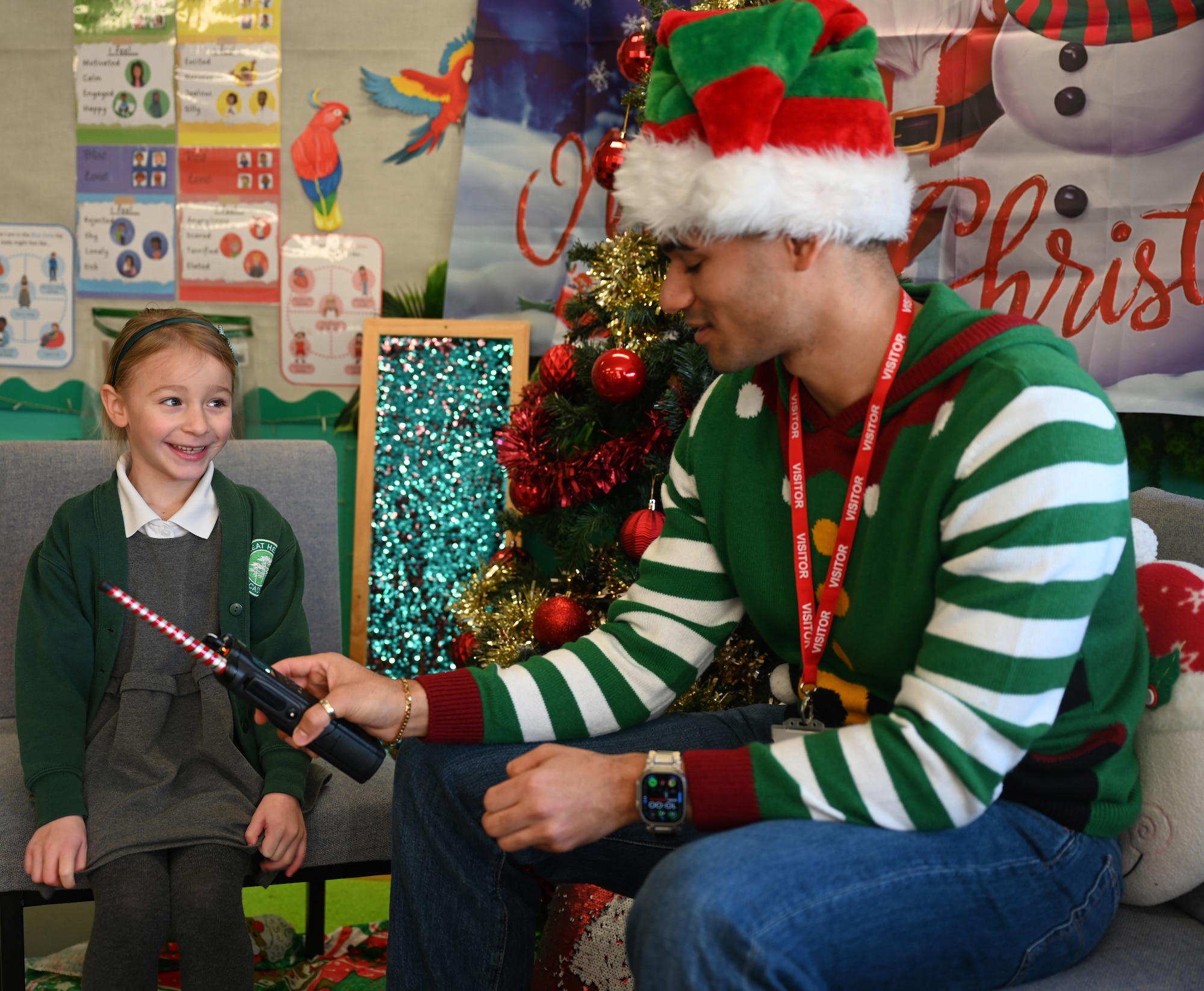 U.S. Air Force Senior Airman Savion Watson, right, 100th Communications Squadron agile combat employment technician, RAF Mildenhall, uses a handheld radio to help a Year-1 child interact with Santa at Great Heath Academy in Mildenhall, Suffolk, Dec. 10, 2025. RAF Mildenhall Airmen regularly volunteer within the local community to strengthen partnerships.  (U.S. Air Force photo by Karen Abeyasekere)
