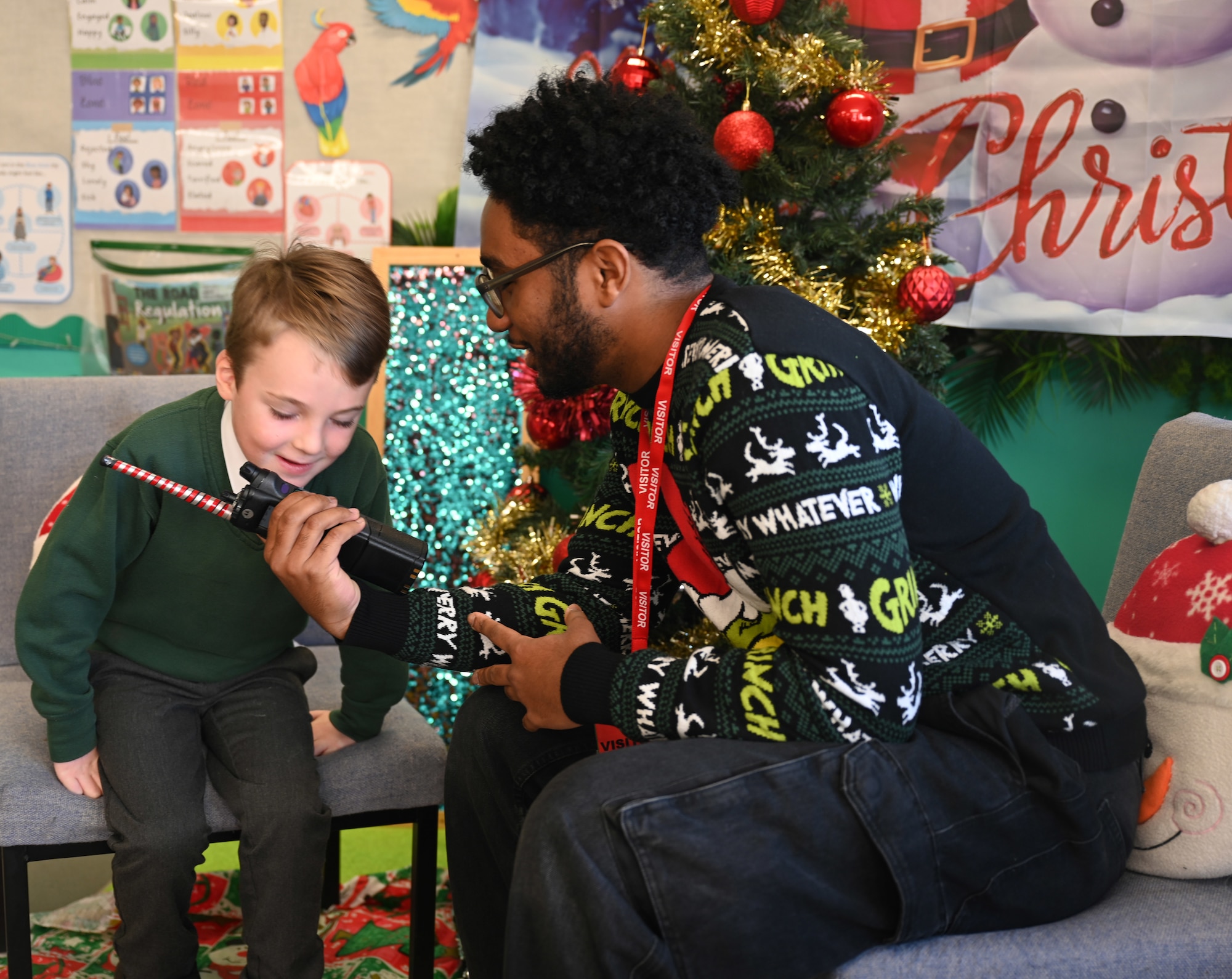 U.S. Air Force Airman 1st Class Jeffrey Favor, right, 100th Communications Squadron radiofrequency transmissions technician, RAF Mildenhall, uses a handheld radio to help a Year-1 child interact with Santa at Great Heath Academy in Mildenhall, Suffolk, Dec. 10, 2025. Three Team Mildenhall members spent time with 47 children and provided them a chance to talk to Santa and make special gift requests before the holidays (U.S. Air Force photo by Karen Abeyasekere)