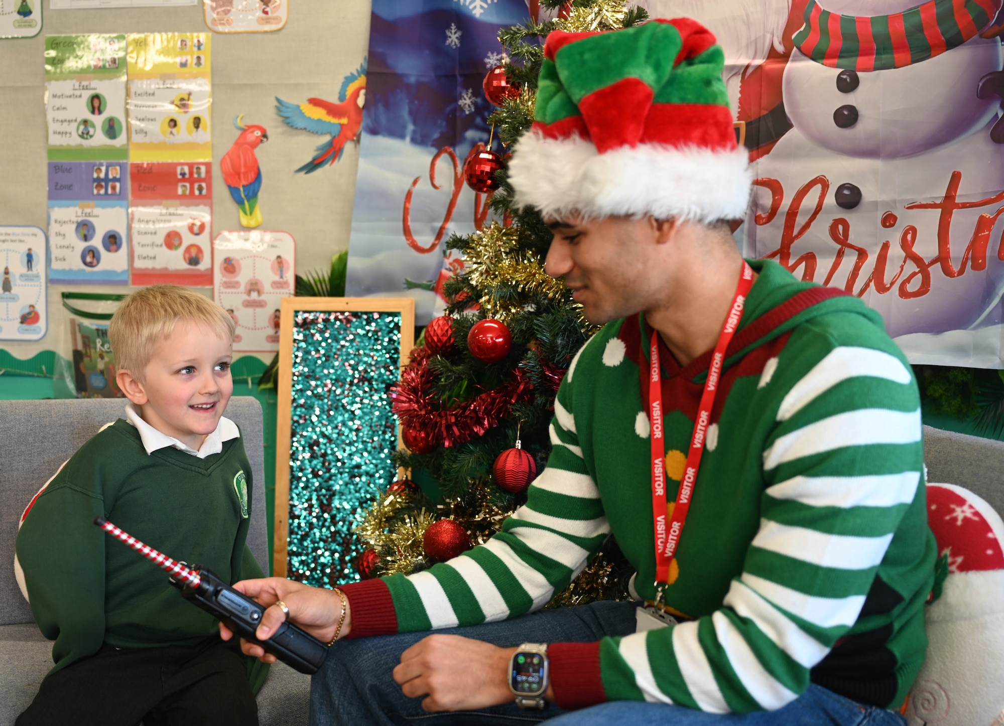U.S. Air Force Senior Airman Savion Watson, right, 100th Communications Squadron agile combat employment technician, RAF Mildenhall, uses a handheld radio to help a Year-1 child interact with Santa at Great Heath Academy in Mildenhall, Suffolk, Dec. 10, 2025. RAF Mildenhall Airmen regularly volunteer within the local community to strengthen partnerships. (U.S. Air Force photo by Karen Abeyasekere)