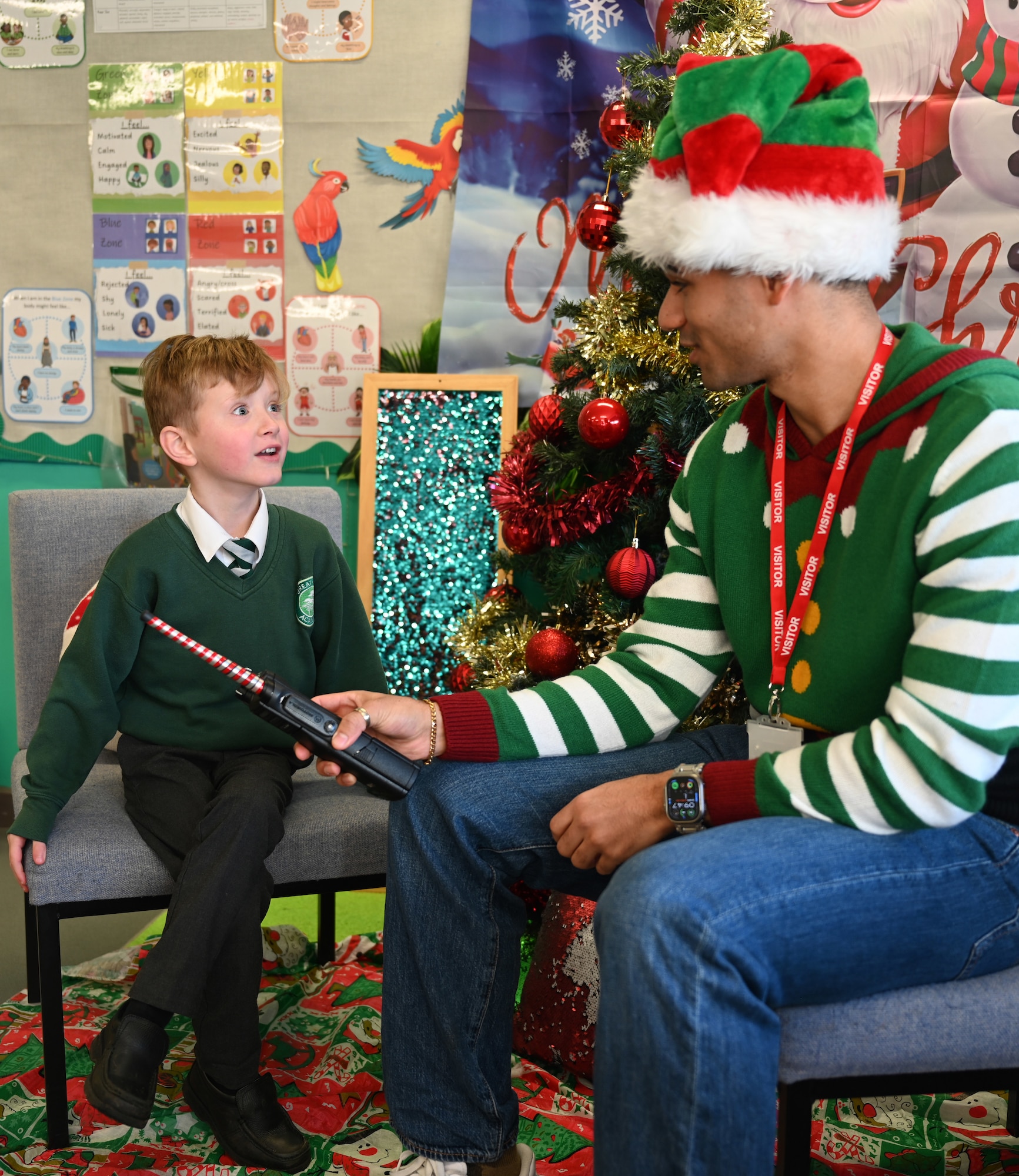 U.S. Air Force Senior Airman Savion Watson, right, 100th Communications Squadron agile combat employment technician, RAF Mildenhall, uses a handheld radio to help a Year-1 child interact with Santa at Great Heath Academy in Mildenhall, Suffolk, Dec. 10, 2025. RAF Mildenhall Airmen regularly volunteer within the local community to strengthen partnerships. (U.S. Air Force photo by Karen Abeyasekere)