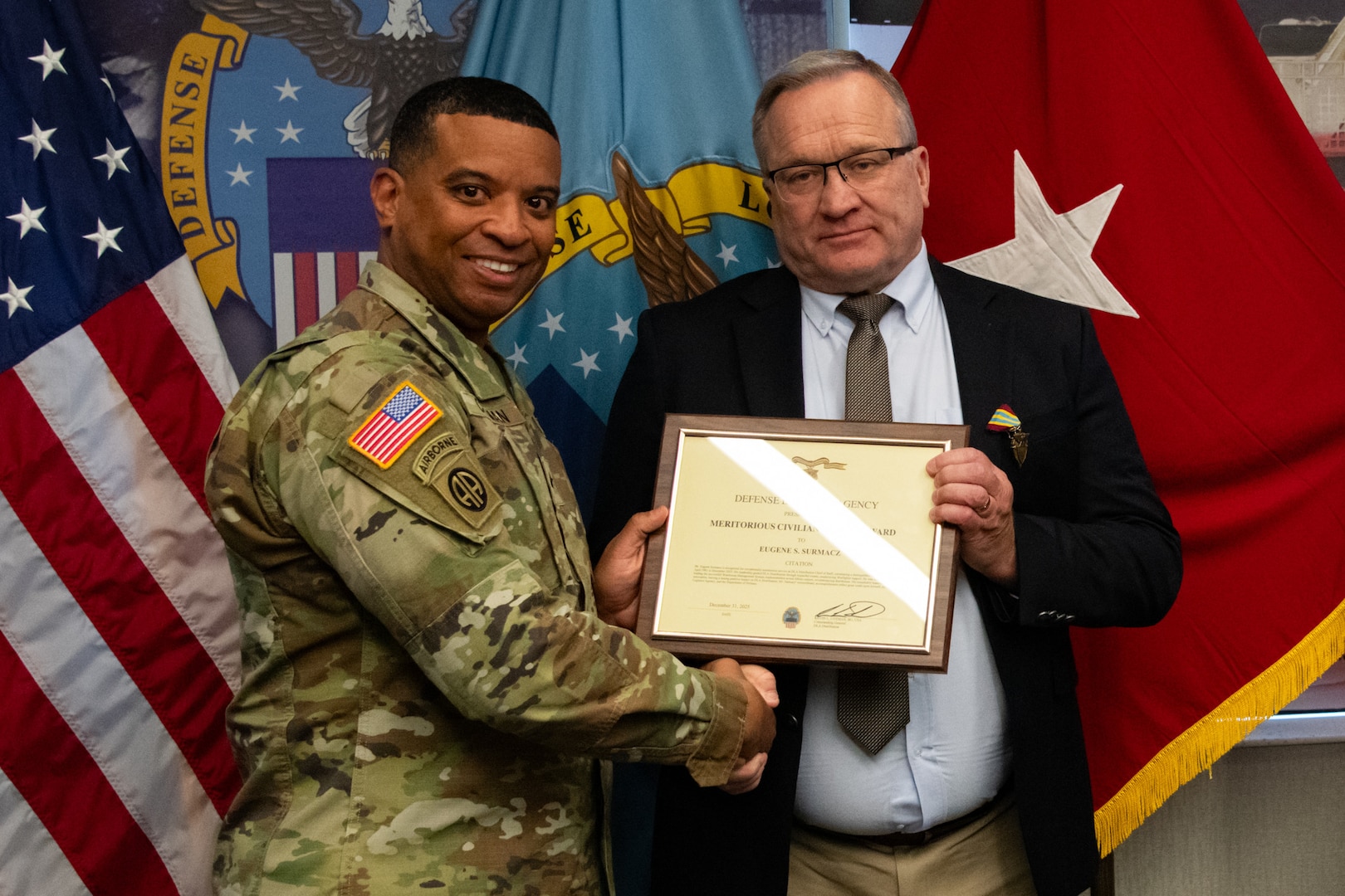 Two people at an award ceremony standing in front of flags.
