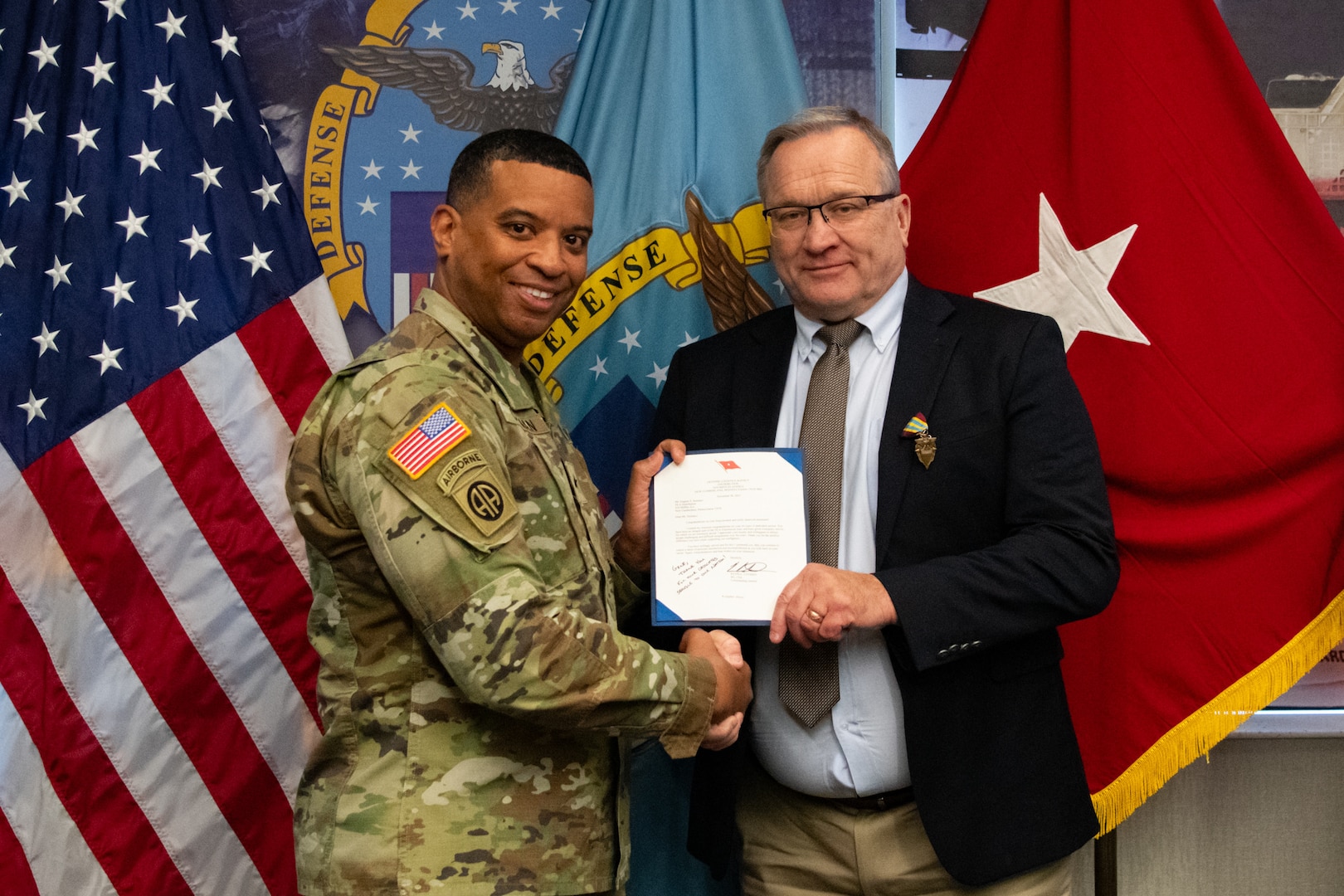 Two people at an award ceremony standing in front of flags.