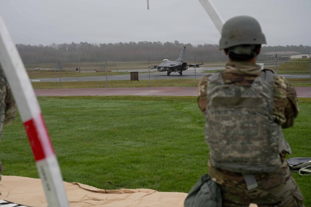 Amn. Charles Peterson, 52nd Civil Engineer Squadron emergency management, observes an F-16 Fighting Falcon during a beddown exercise at Spangdahlem Air Base, Germany, Dec. 5th, 2025. Tent assembly is a vital skill as it directly supports deployment and contingency operations. (U.S. Air Force photo by Senior Airman Darius Frazier)