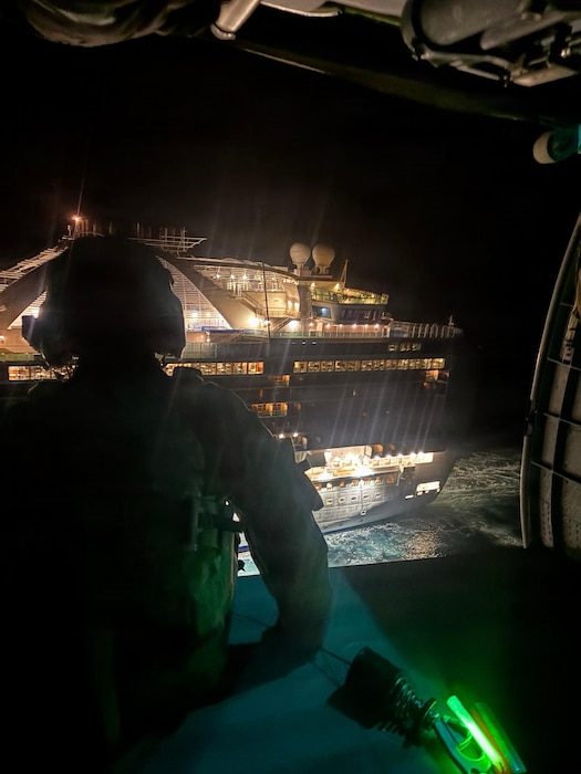 An Airman sits on the open ramp of a helicopter, looking at a cruise ship illuminated at night.