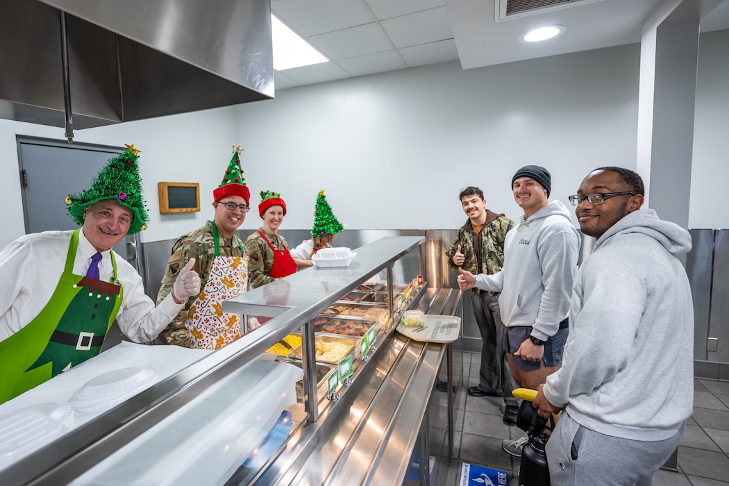 Volunteers, wearing holiday themed hats and aprons, and three young men, pose for a group photo standing at a stainless-steel serving counter