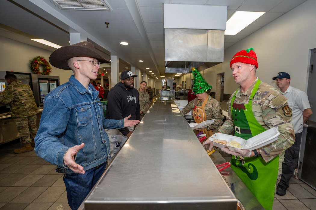 A young man, wearing a dark cowboy hat, denim jacket and pants, smiles as a man, wearing camouflage military uniform, red elf hat, and green elf apron, holds a Styrofoam to-go box. A stainless-steel counter runs between them