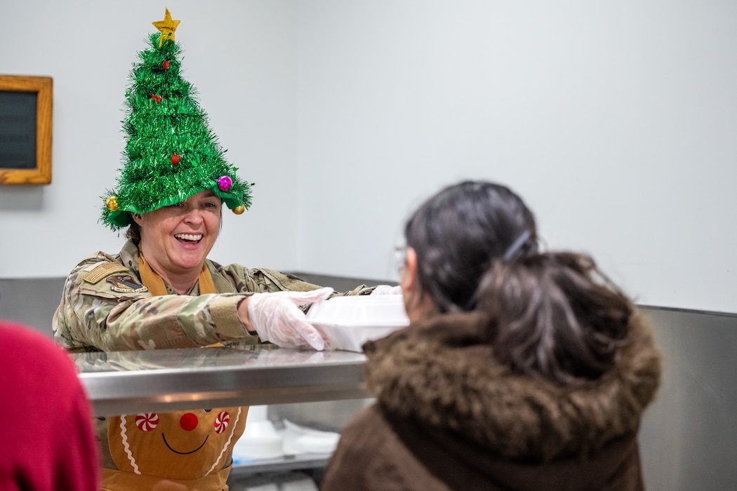 A woman, wearing a camouflage military uniform with a sparkly Christmas tree cone hat and gingerbread apron, smiles as she hands a to-go dinner box to a woman with dark drown winter coat