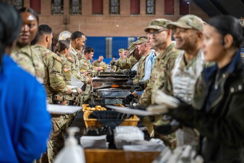 Men and women wearing military camouflage uniforms stand in two lines in front of a food service area for a Thanksgiving meal.