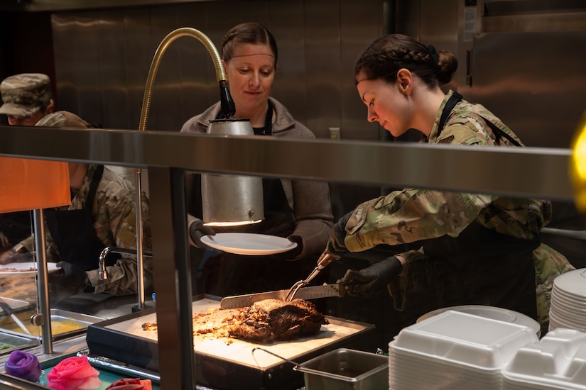 A woman wearing a military camouflage uniform and apron slices a roast while another woman dressed similarly watches.