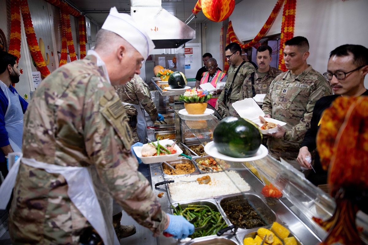 Men dressed in military camouflage uniforms and civilian attire stand in line while other men wearing aprons and chef hats along with military camouflage uniforms serve food.