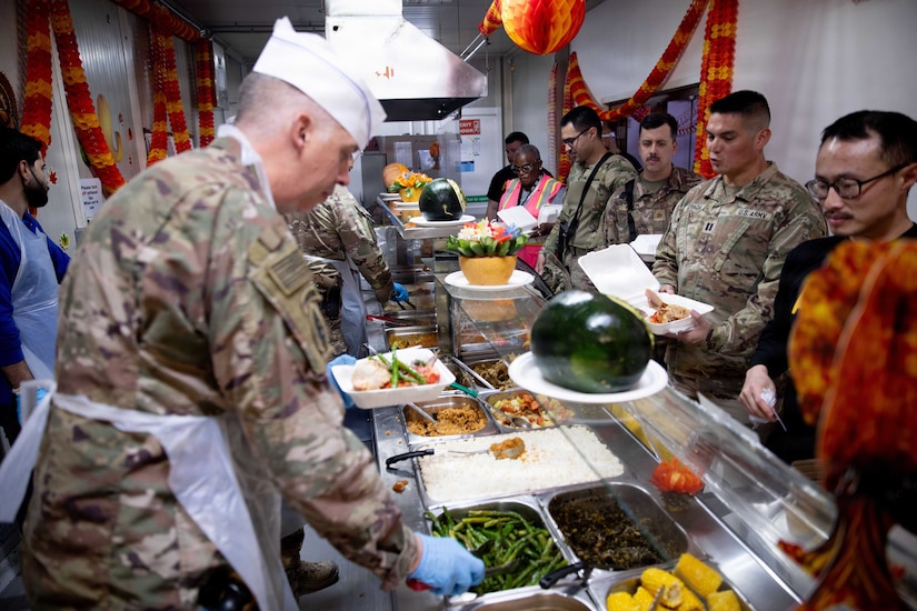 Men dressed in military camouflage uniforms and civilian attire stand in line while other men wearing aprons and chef hats along with military camouflage uniforms serve food.