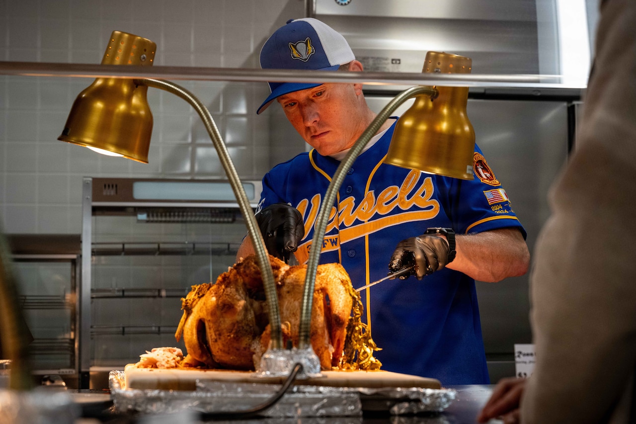 A man wearing a sports shirt and a baseball cap cuts a turkey for serving.
