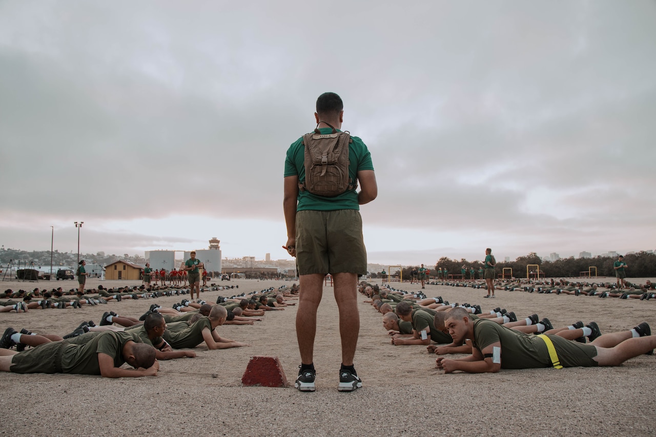 A Marine in athletic wear stands and monitors a group of Marines in similar attire who are lying on the ground while doing physical fitness.