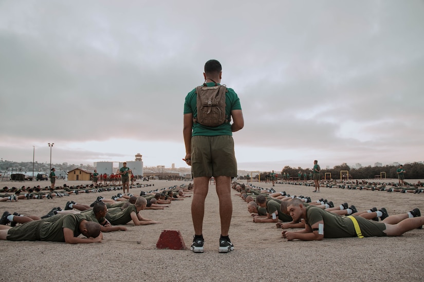 A Marine in athletic wear stands and monitors a group of Marines in similar attire who are lying on the ground while doing physical fitness.
