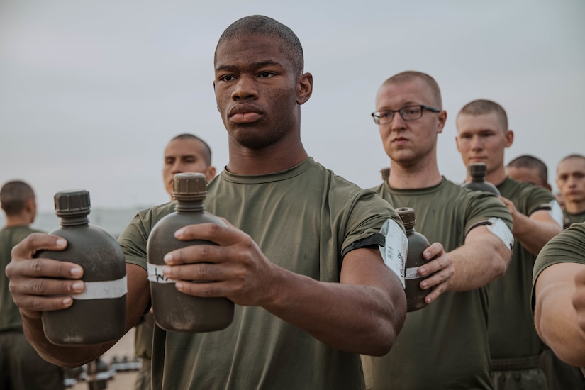 Marines in athletic wear each hold two canteens while standing in formation during a physical fitness test.