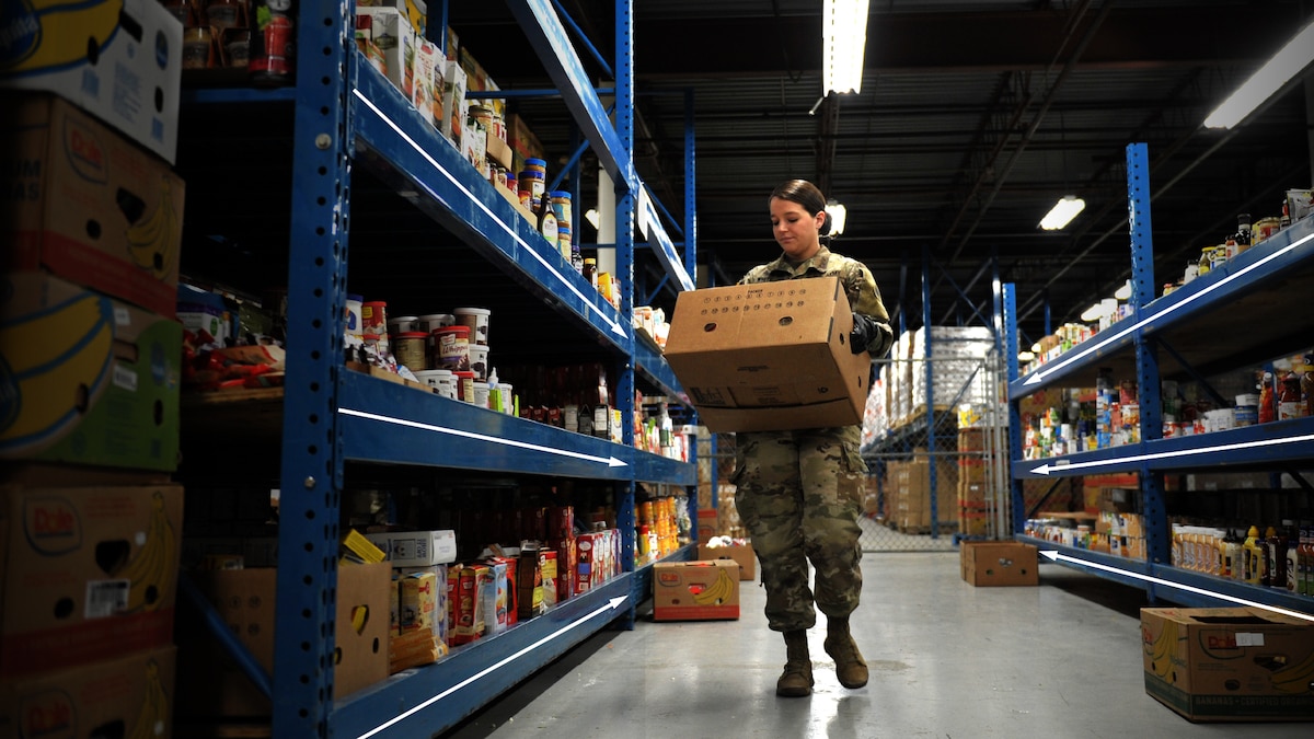 A uniformed service member in a storage room carries a box of supplies toward the camera between two tall parallel shelving units.
