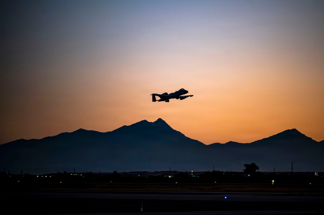 An aircraft flies in dark blue, orangish sky over the silhouette of a mountain.