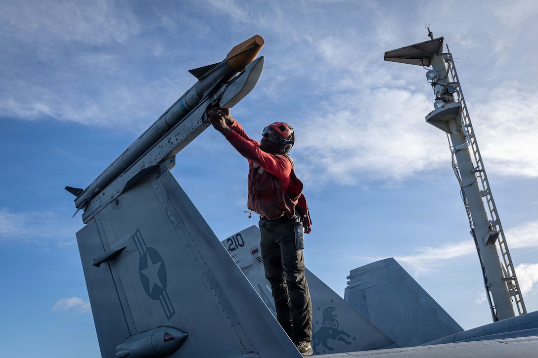 A sailor works on ordnance on an aircraft aboard a ship at sea against a cloudy, blue sky.