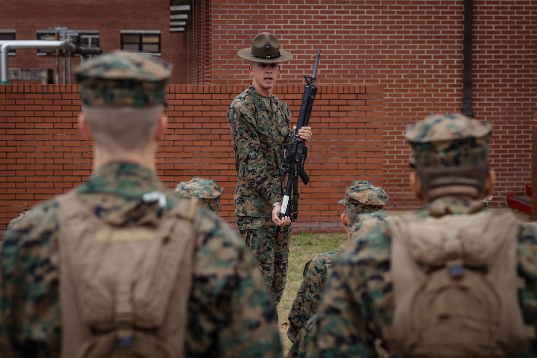 A Marine instructs recruits while holding a weapon as they stand in front of a brick building.