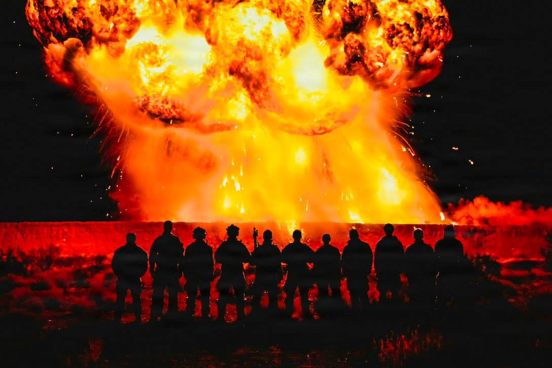 Eleven explosive ordnance disposal technicians pose for a photo in silhouette in the foreground as an explosion creates a massive fireball in the background.