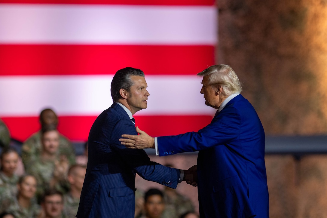 President Donald J. Trump and Defense Secretary Pete Hegseth shake hands in front of a seated troop audience and U.S. flag.