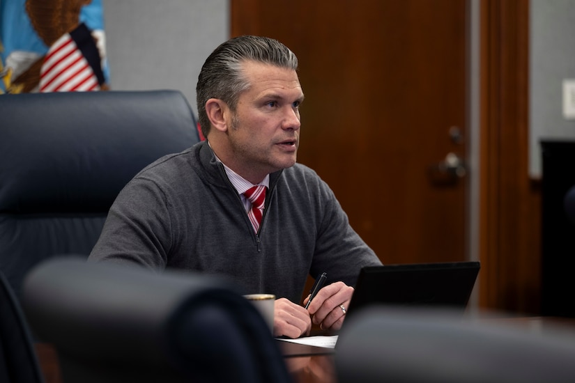 A man in a sweater and tie sits at a table in a room, looking to the right of the screen.