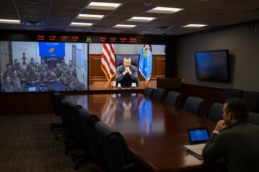 A man in a sweater and tie sits at a table in a room, smiling as he looks to the right of the screen.