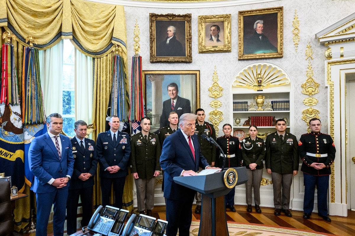A man in business attire stands behind a lectern in an ornate room. Behind him stand a dozen service members in formal military uniform, as well as a man in business attire.