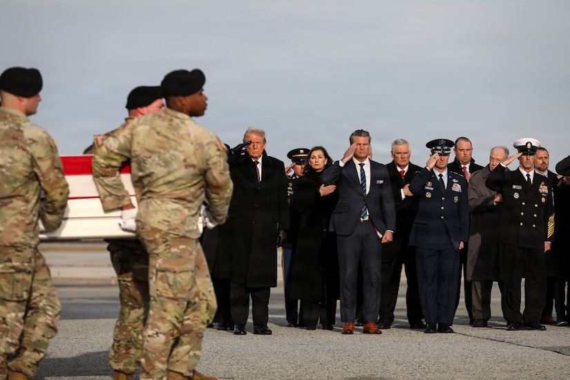 Uniformed personnel carry a casket draped with an American flag. In the rear, people in business attire and formal military uniforms either salute or place their hands over their hearts.