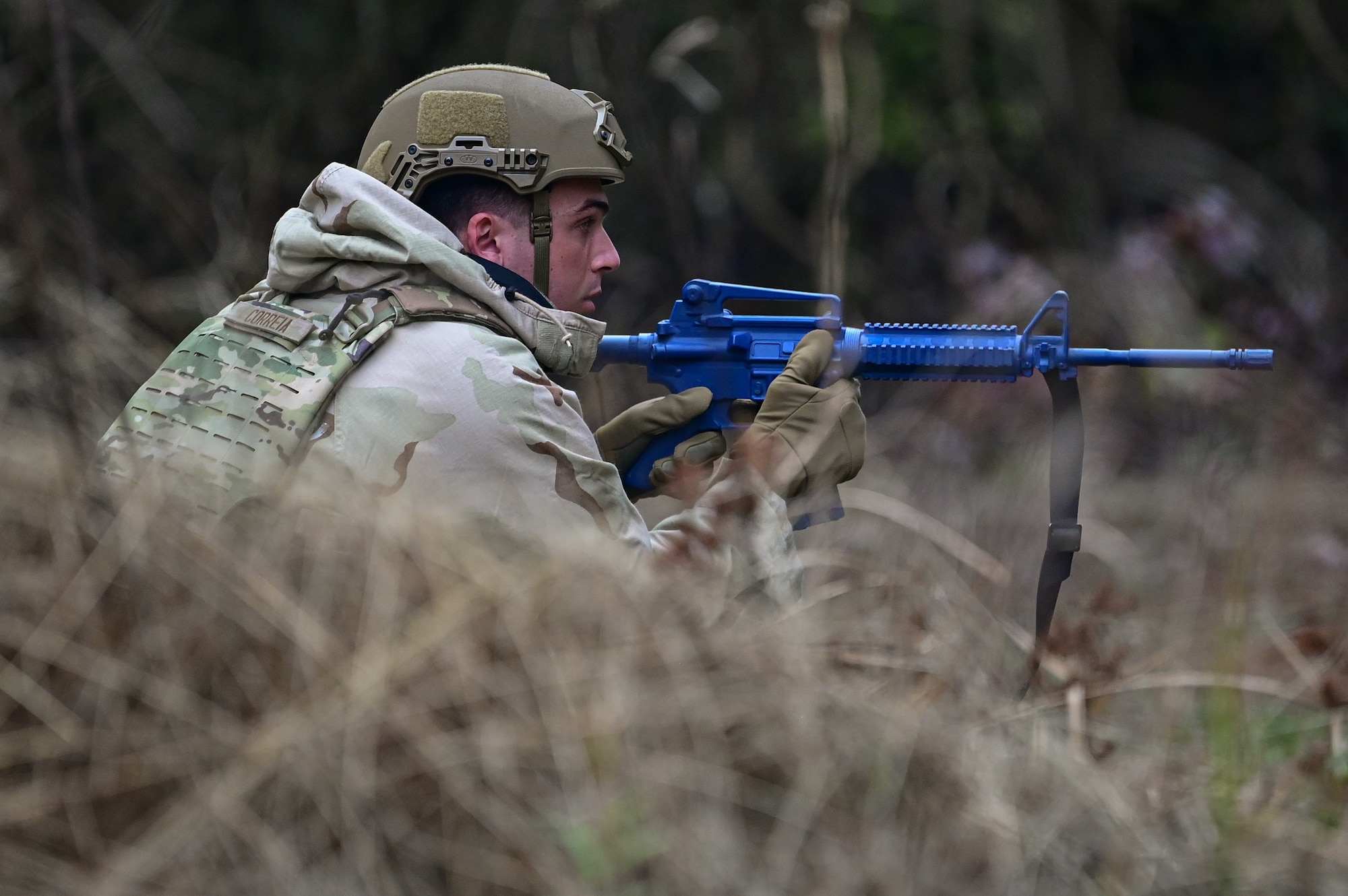 An Airman kneels in the brush with a blue training assault rifle during an exercise.